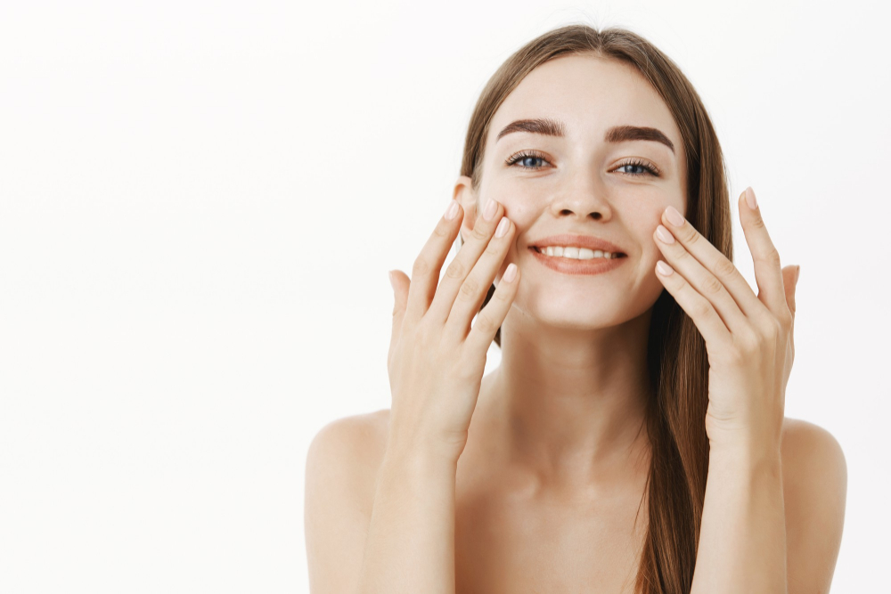 charming-relaxed-gentle-young-woman-making-cosmetological-procedure-applying-facial-cream-face-with-fingers-smiling-broadly-feeling-perfect-taking-care-skin